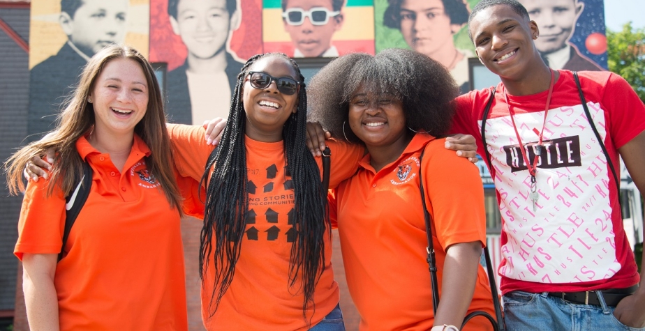 Smiling students in orange T-shirts pose in front of an Elwood Village banner