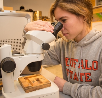 Student wearing a Buffalo State sweatshirt and looking into a microscope